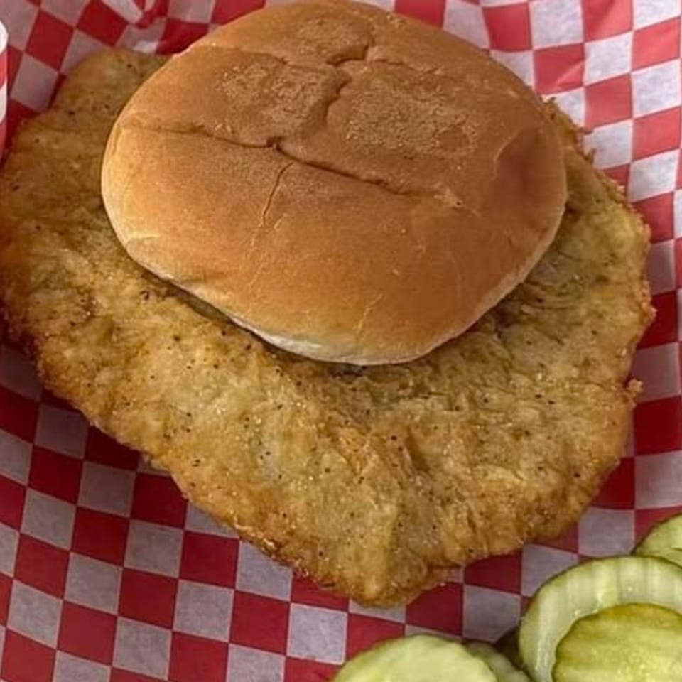 A large fried chicken sandwich on a bun, served with pickles in a red and white checkered basket.